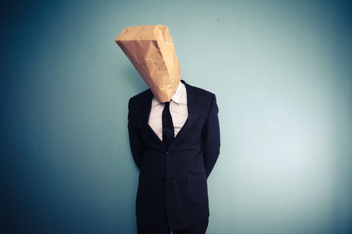 Person in a business suit with a brown paper bag over their head.